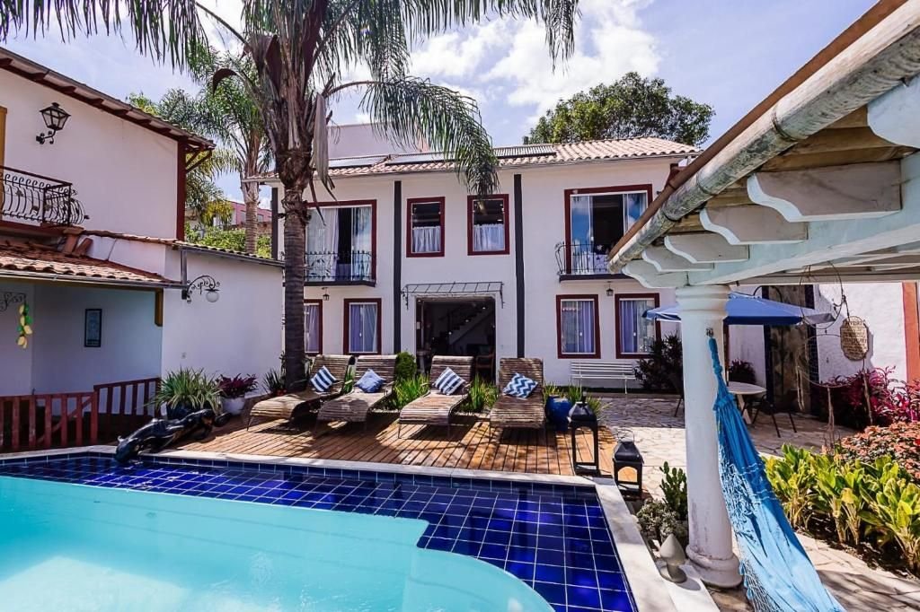 Piscina de pastilhas azuis com deck de madeira, espreguiçadeiras, sede da pousada e pergolado com rede